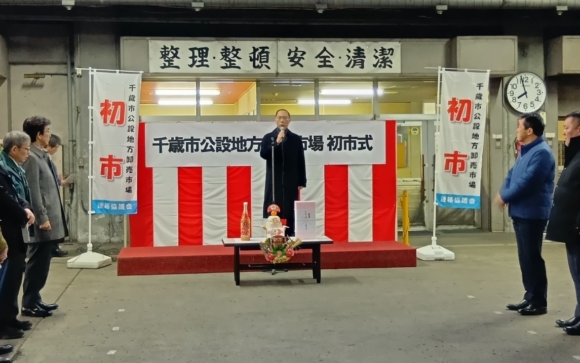 紅白幕の舞台上で挨拶をする市長とその両脇で市場関係者が挨拶を聞いている写真