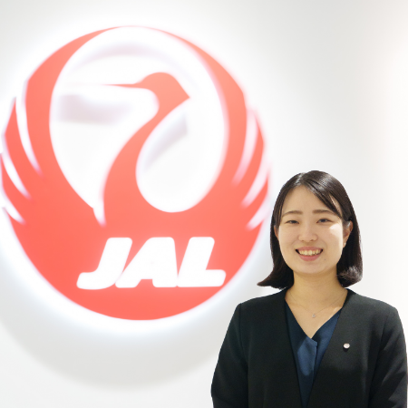 壁に飾られた大きなJALのロゴの前で微笑む伊橋さんの写真